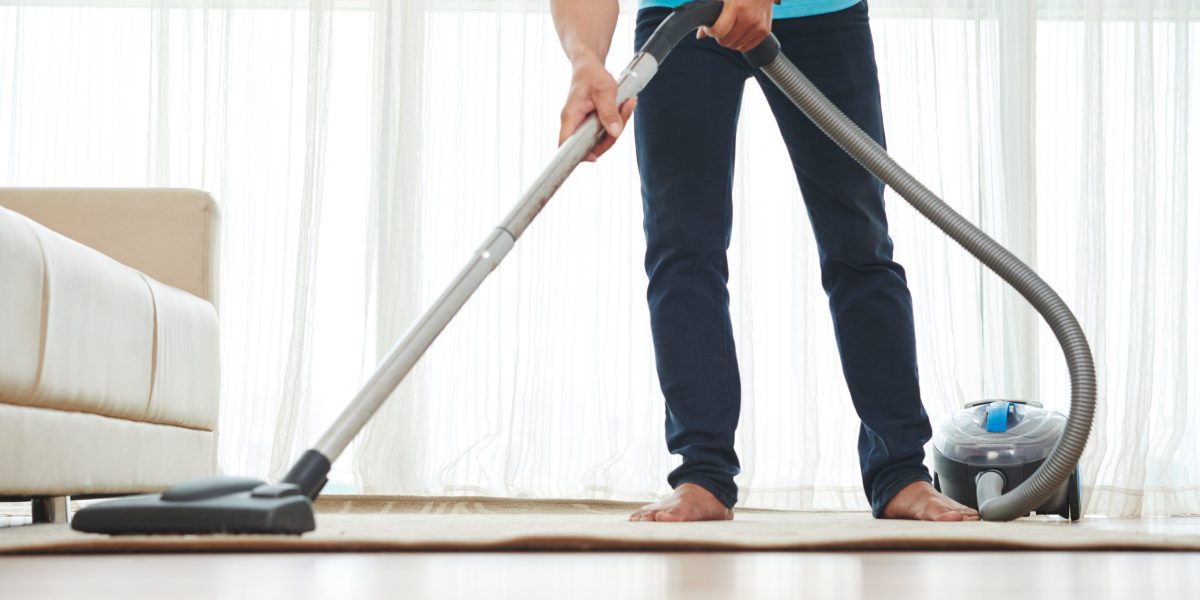 Unrecognizable person using a canister vacuum to clean a beige carpet in a bright living room